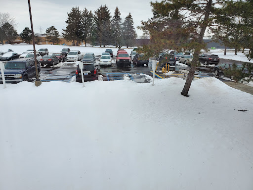 A snowy parking lot with several vehicles visible, surrounded by trees under a cloudy sky.