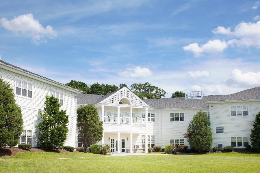 A residential building with a white exterior, surrounded by trees and a well-maintained lawn under a blue sky with clouds.
