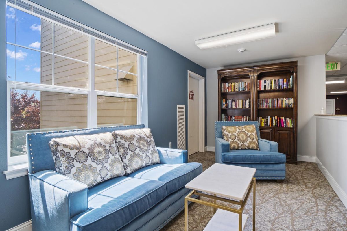 A cozy living room with a blue sofa and armchair, a bookshelf filled with books, and a window allowing natural light in.
