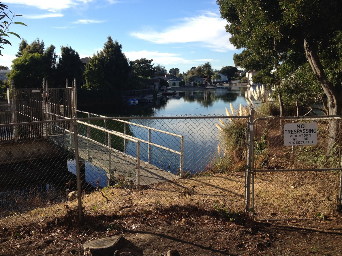 A view of a serene lake behind a chain-link fence, surrounded by trees and under a clear sky.