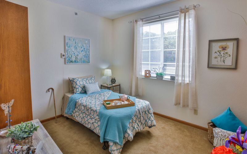 A cozy bedroom featuring a single bed with blue and white bedding, a bedside lamp, and a window allowing natural light to enter.