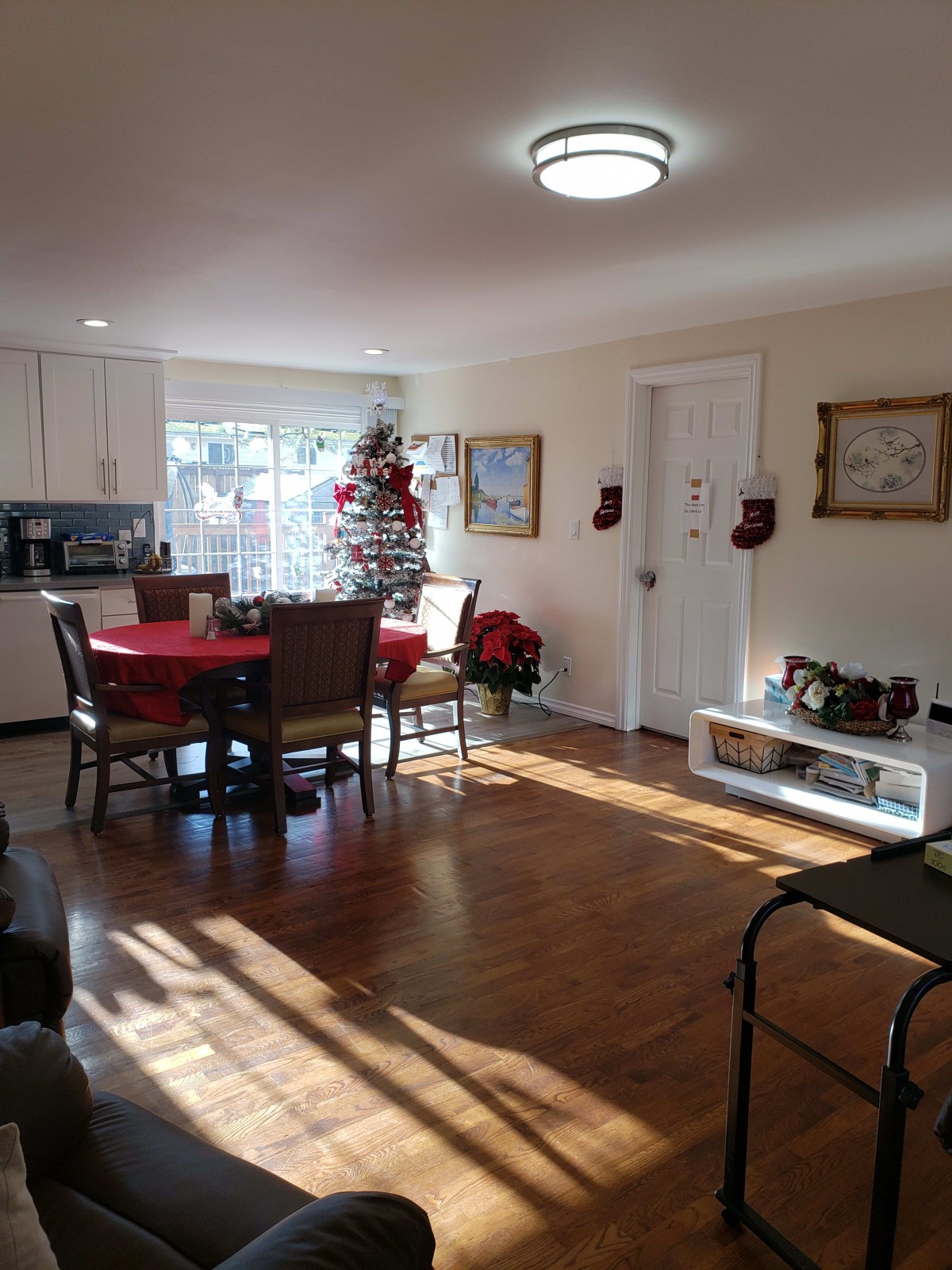 A cozy living and dining space decorated for Christmas with a tree and festive red accents, illuminated by sunlight.