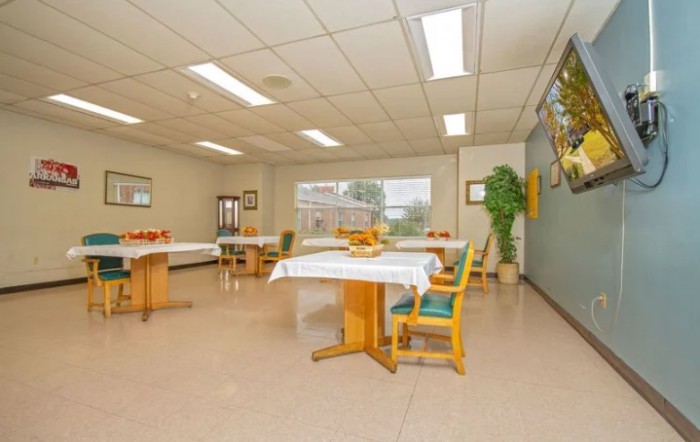 A spacious dining room with neatly arranged tables and chairs, well-lit by overhead fluorescent lights. Large windows allow natural light in, complemented by potted plants for a touch of greenery.
