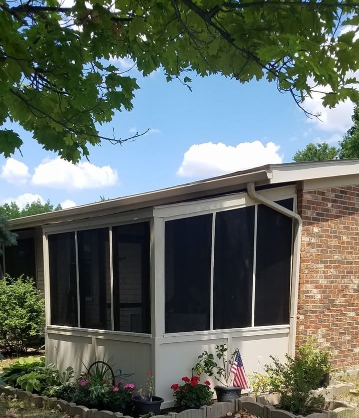 A house with a screened porch and a brick exterior, surrounded by trees and a garden under a blue sky.