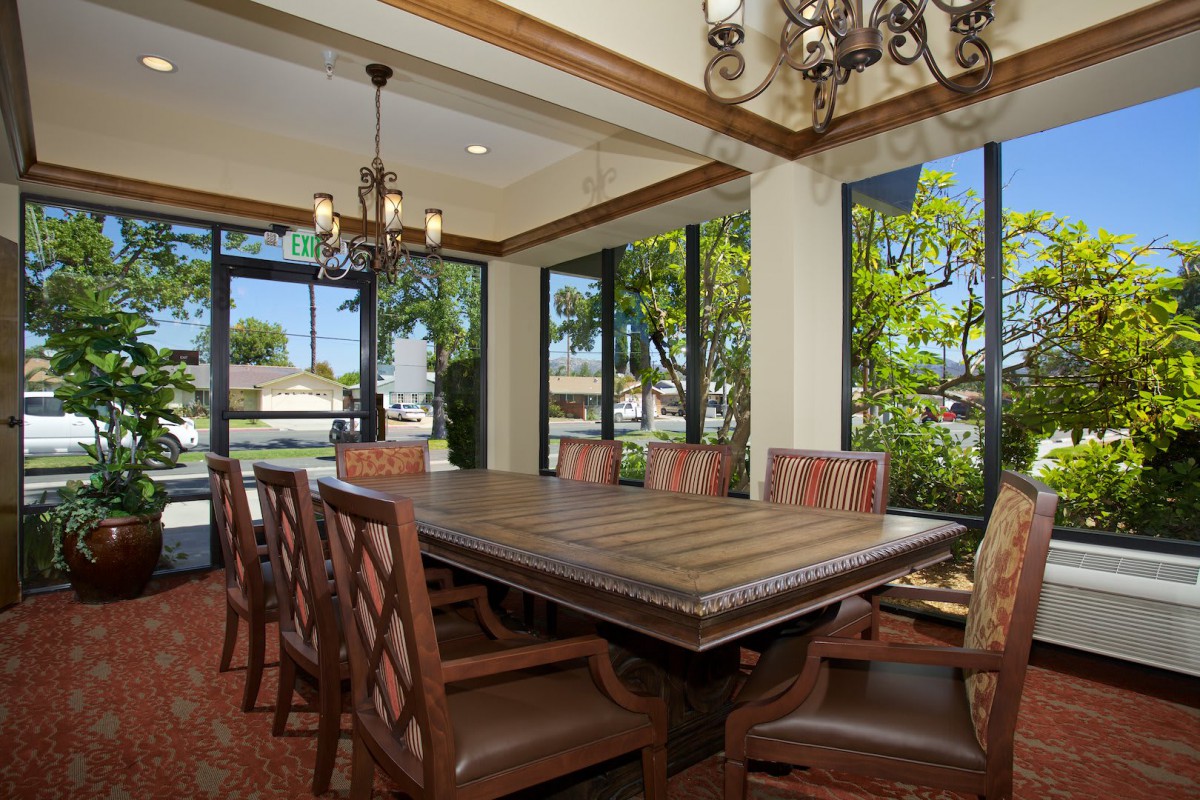 A spacious dining room with a large wooden table, surrounded by chairs and illuminated by chandeliers, featuring large windows that offer ample natural light.
