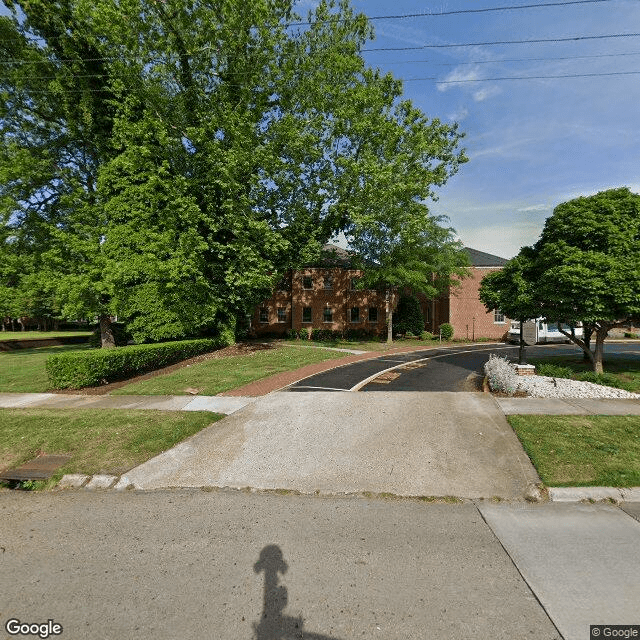 A suburban driveway leading to a brick building surrounded by trees and a well-maintained lawn.