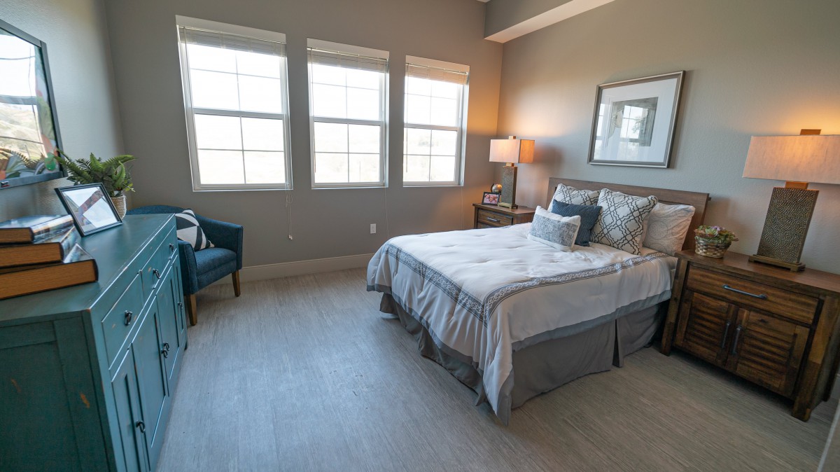A cozy bedroom with a neatly made bed, a lamp on the side table, and three large windows allowing natural light to fill the room.