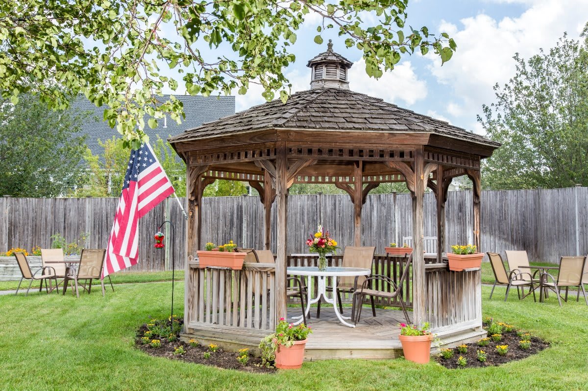 A charming wooden gazebo in a garden setting, adorned with potted plants and an American flag nearby.