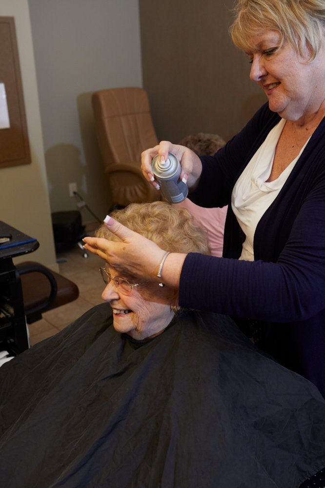 An elderly woman getting her hair styled at a salon, smiling as the hairdresser applies hairspray.