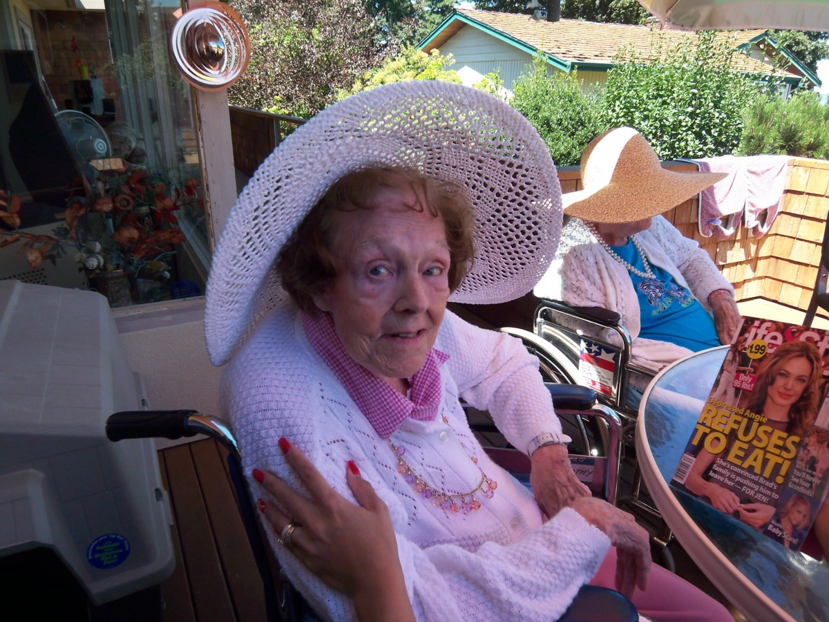 An elderly woman wearing a sun hat sits outdoors in a wheelchair.