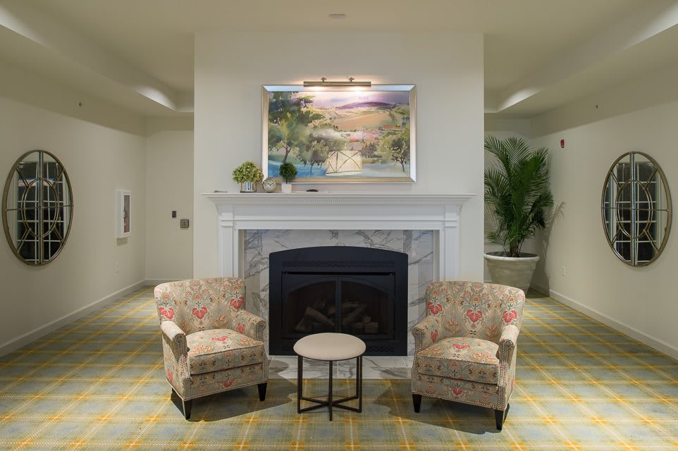 A cozy living room setup featuring a fireplace flanked by two floral-patterned chairs and a small round table, with artwork and greenery adding to the ambiance.