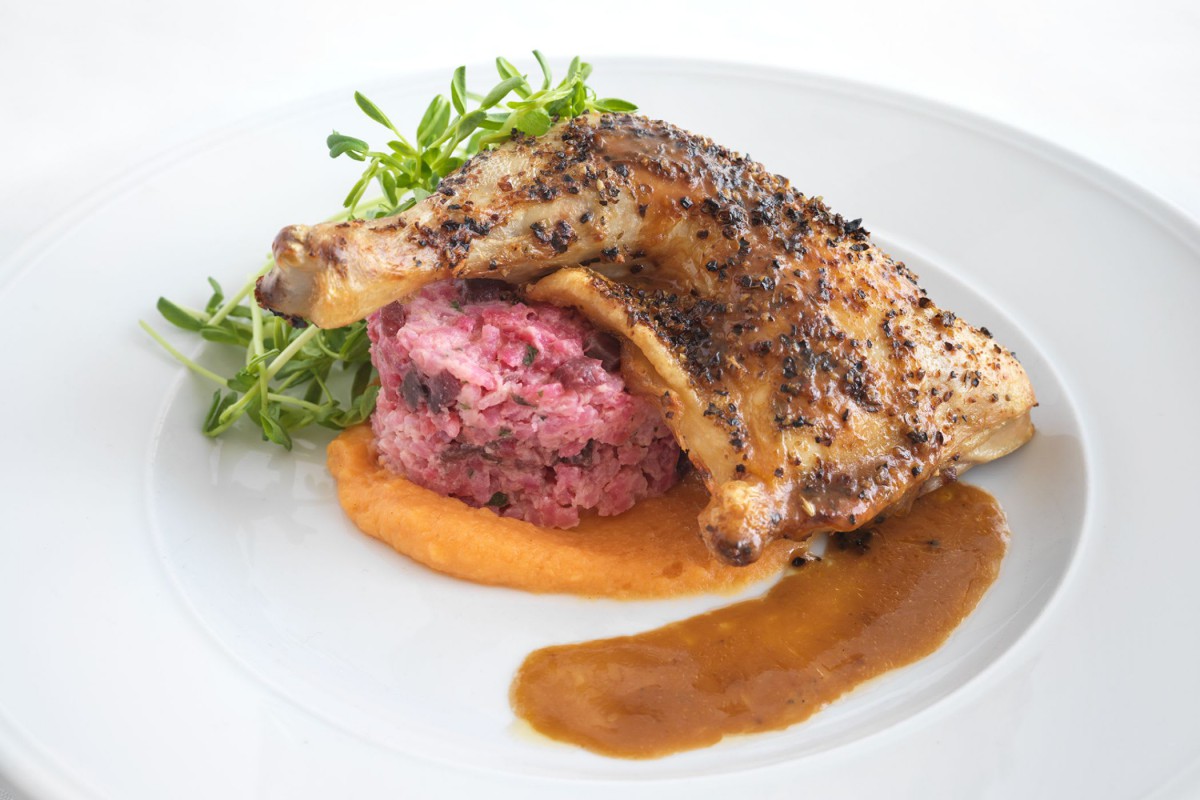A gourmet dish featuring a seasoned chicken thigh atop a bed of pink mashed potatoes, garnished with fresh herbs and accompanied by a rich sauce.