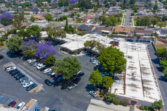 An aerial view of a suburban area with buildings, a parking lot, and several parked cars surrounded by trees.
