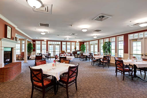 A spacious dining room with neatly arranged tables and chairs, well-lit by ceiling lights and surrounded by large windows.