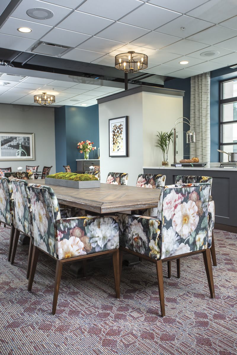 A modern dining room with a large wooden table surrounded by floral-patterned chairs, illuminated by chandeliers and framed by large windows.