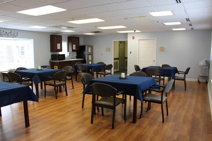 A dining area with multiple tables covered in blue tablecloths and chairs, featuring a kitchen setup and wooden flooring.