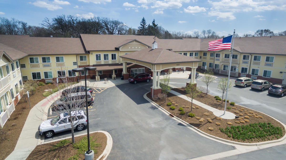 A two-story building with a driveway and parked cars, surrounded by landscaped grounds under a partly cloudy sky.