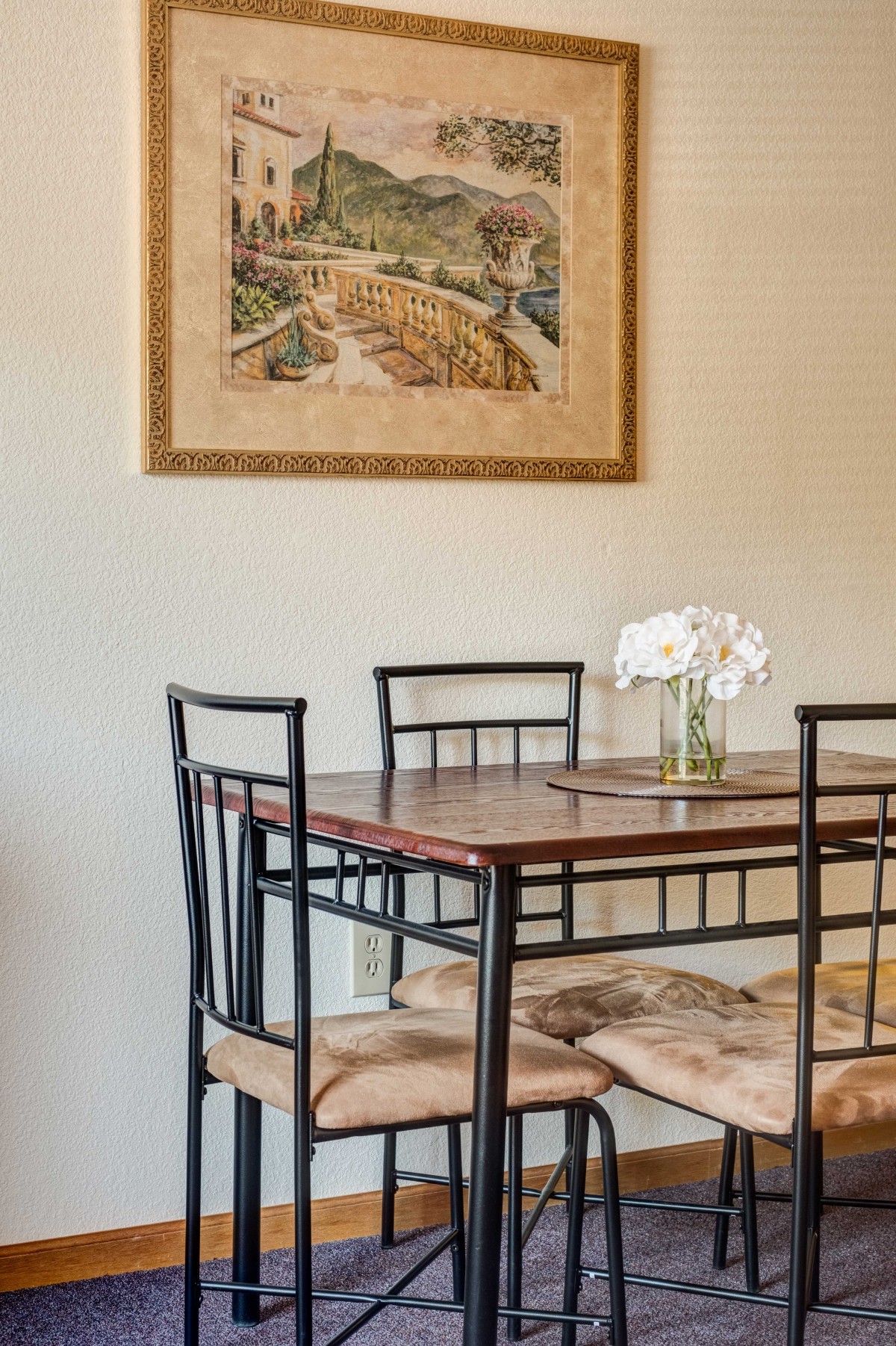 A cozy dining area with a table set for four, featuring a vase of flowers and wall art.