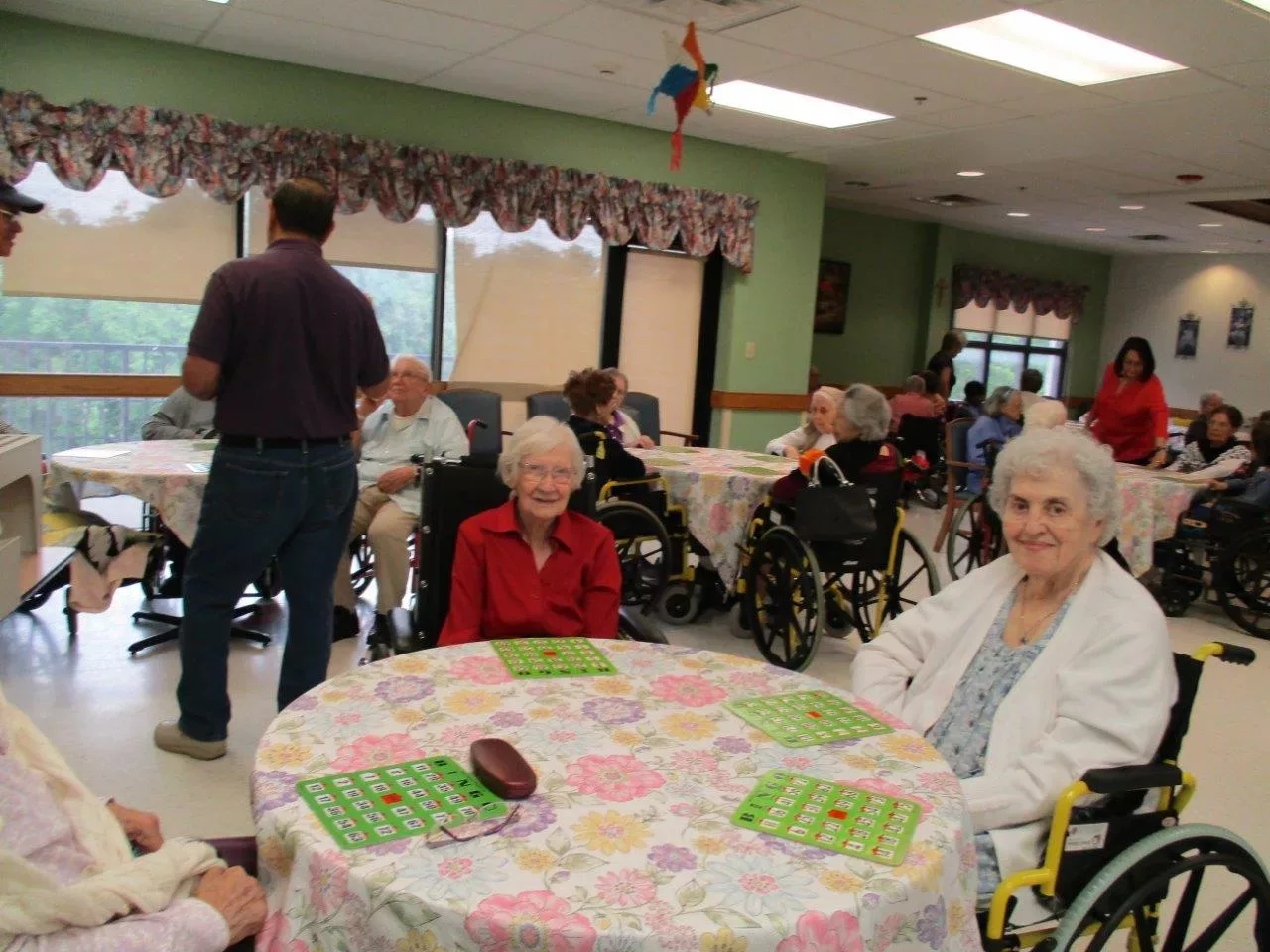 Elderly individuals gathering in a community room, some seated in wheelchairs and playing bingo.
