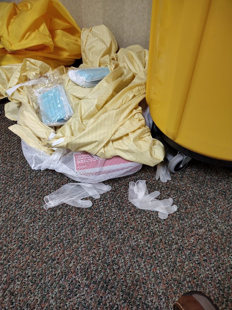 Discarded medical waste including gloves, face masks, and gowns near a trash bin on a carpeted floor.