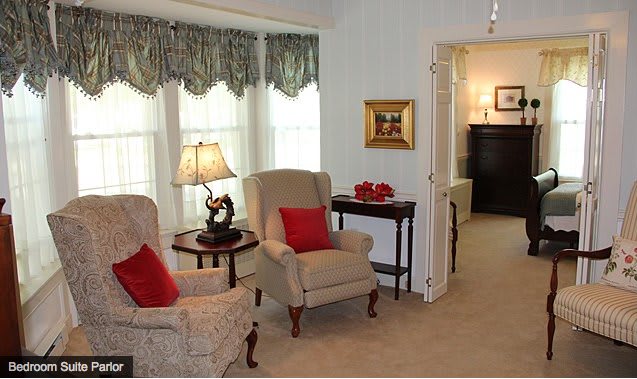 A cozy living room with armchairs, a lamp, and decorative pillows against a backdrop of large windows with curtains.