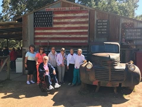 A group of elderly individuals poses in front of a rustic building featuring a large American flag and an old, rusted truck.