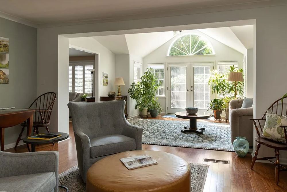 A cozy living room with comfortable armchairs, a round coffee table, and large windows allowing ample natural light. The space is decorated with plants and features a hardwood floor.