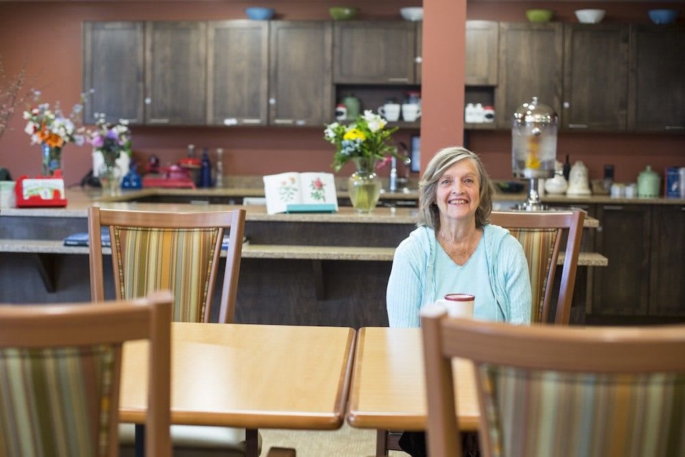 A woman sits smiling in a cozy cafe with a kitchen area in the background.