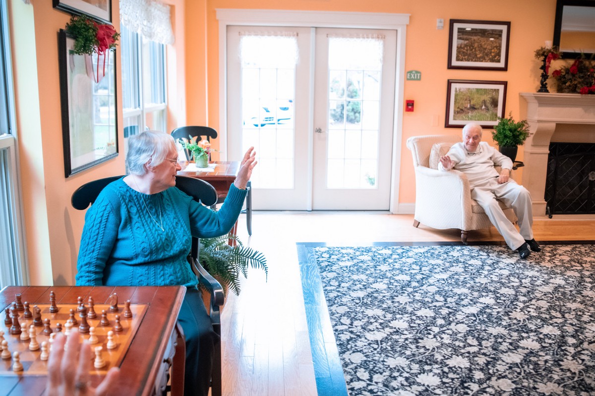 Two seniors interacting in a cozy room, with a chessboard nearby.