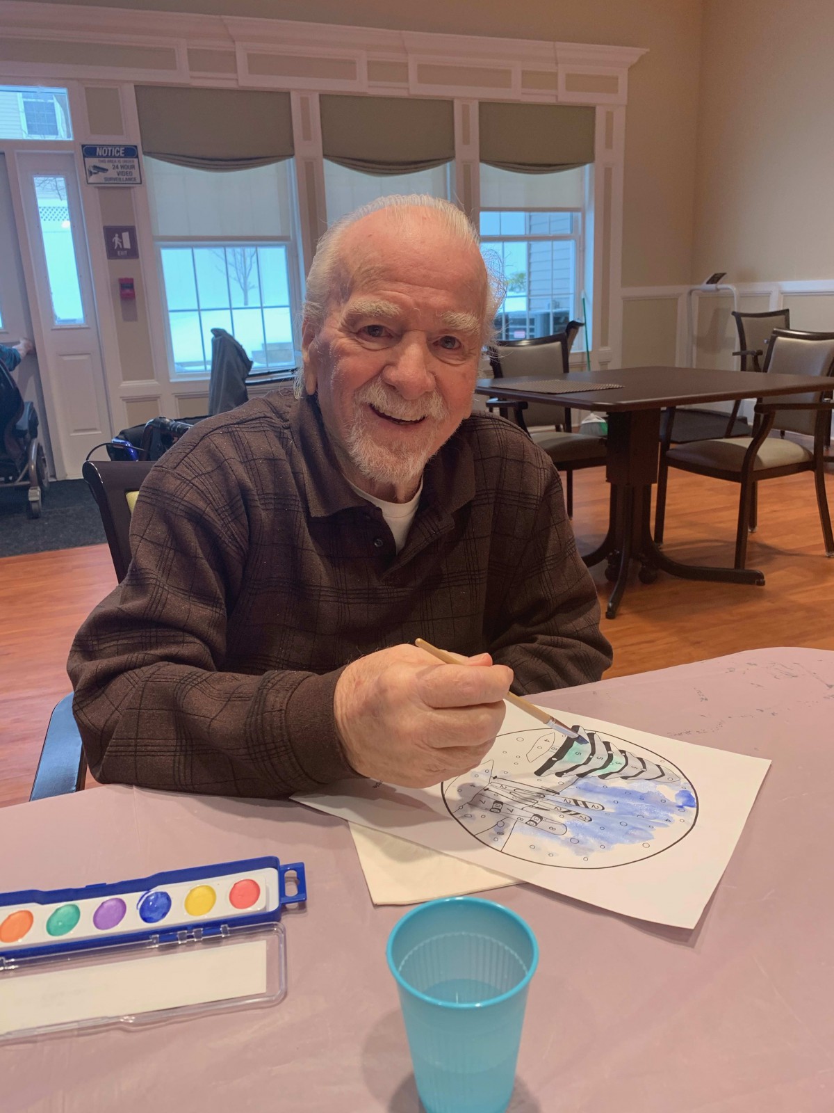 An elderly man smiling while engaging in a painting activity indoors.