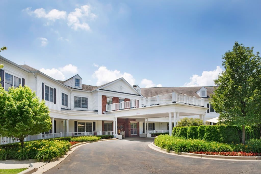 A large white residential building with a driveway, surrounded by landscaped greenery and under a partly cloudy sky.