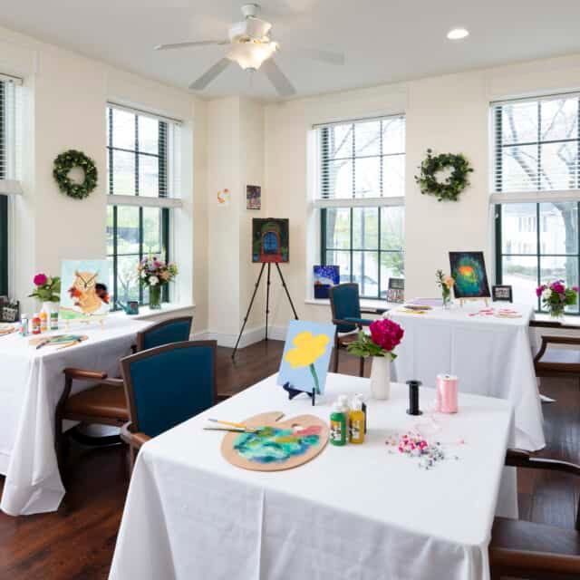 A bright art studio with tables set up for painting, featuring easels and art supplies.