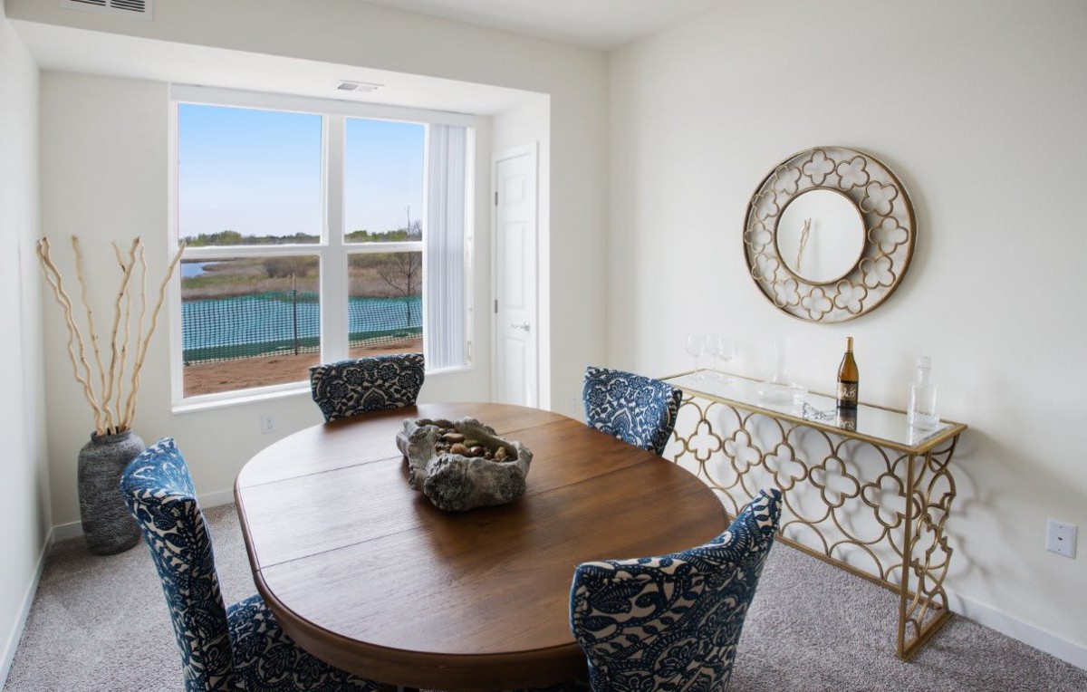 A bright dining room with a round wooden table and patterned chairs overlooks a lake through a large window.