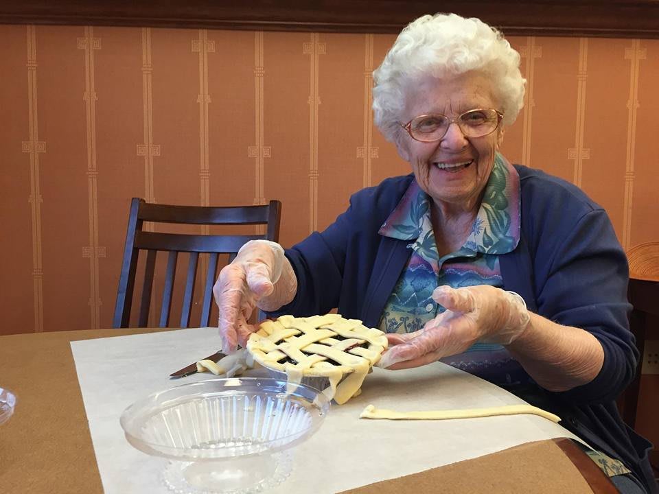 An elderly woman happily preparing a pie with a lattice crust in a kitchen setting.