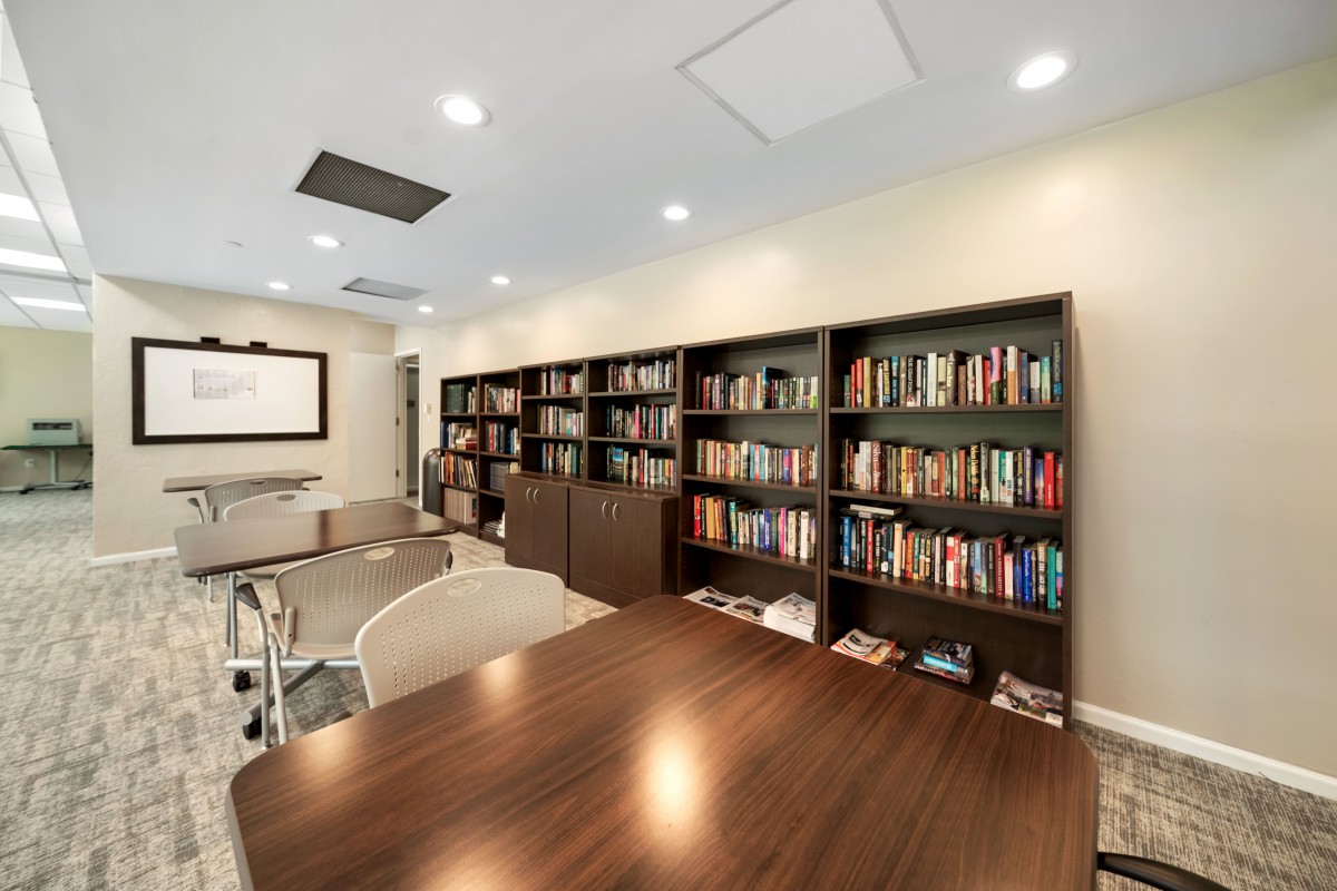 A modern library or conference room with bookshelves filled with books, a large wooden table, and chairs for meetings.