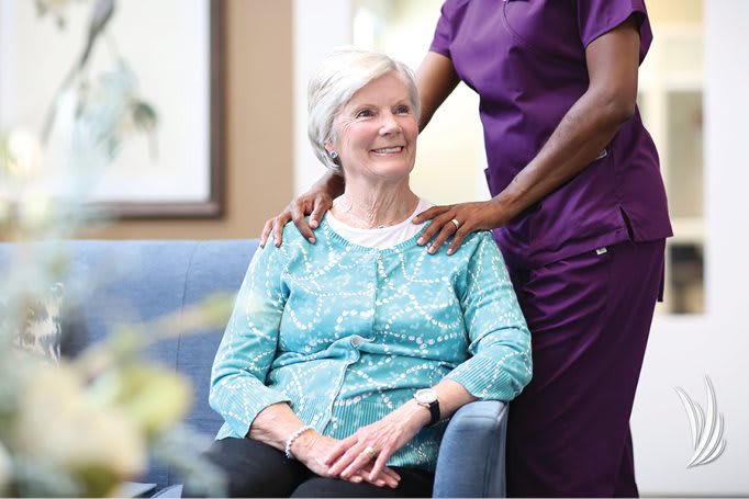 A senior woman sits comfortably while a healthcare professional provides support.