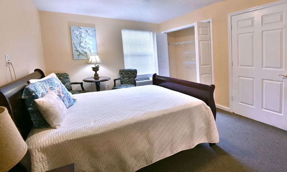 A cozy bedroom with a neatly made bed, two chairs, and a lamp by the window.