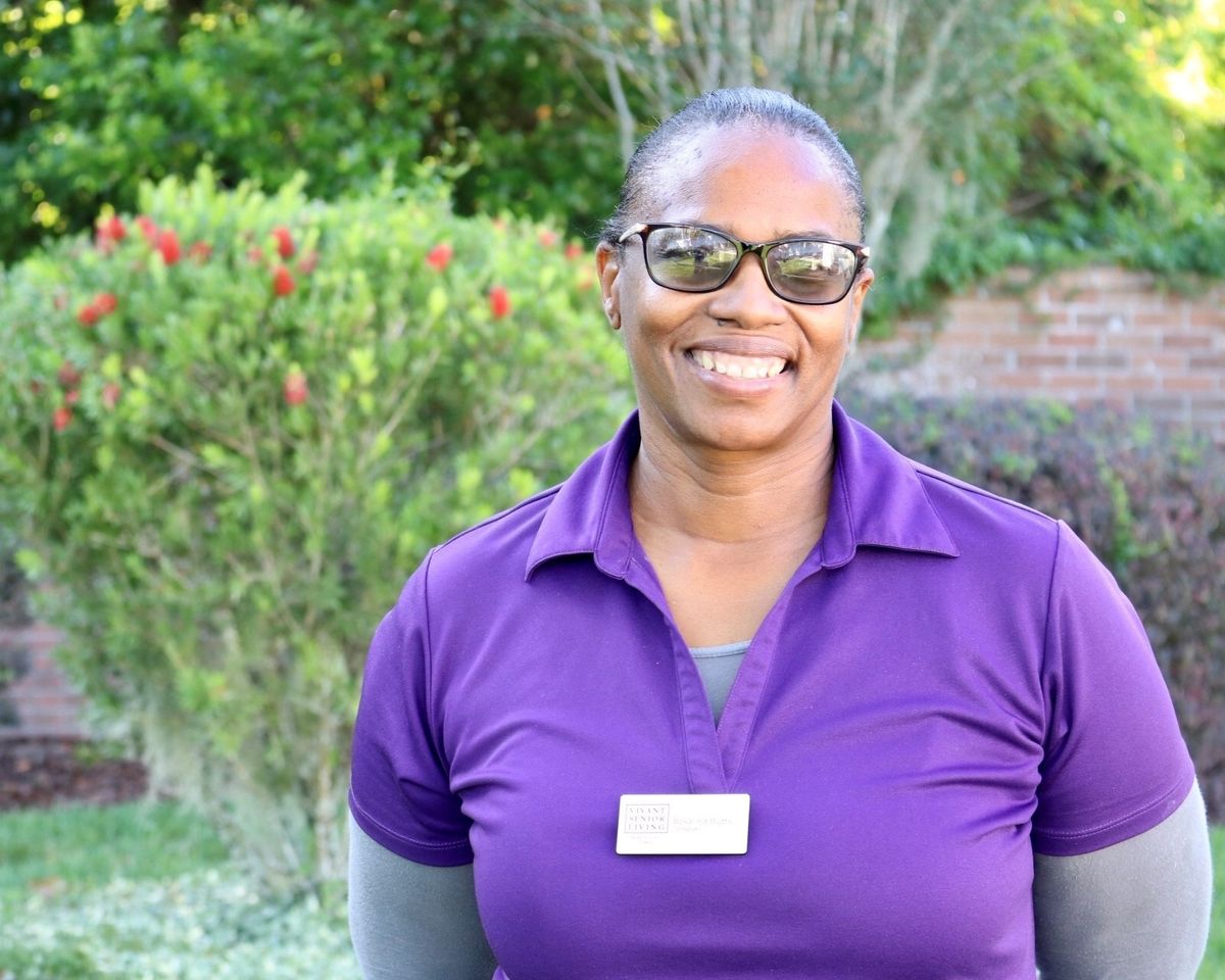 A woman wearing glasses and a purple shirt is smiling outdoors.