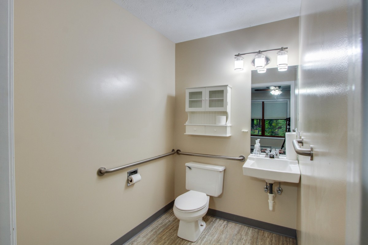 A clean and accessible bathroom featuring a toilet, sink with mirror, and wall-mounted cabinet equipped with grab bars.