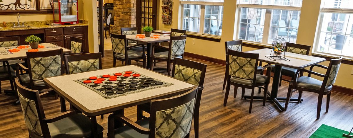 A cozy dining area featuring several tables and chairs with decorative table settings, set against a wooden floor and natural light streaming in through the windows.