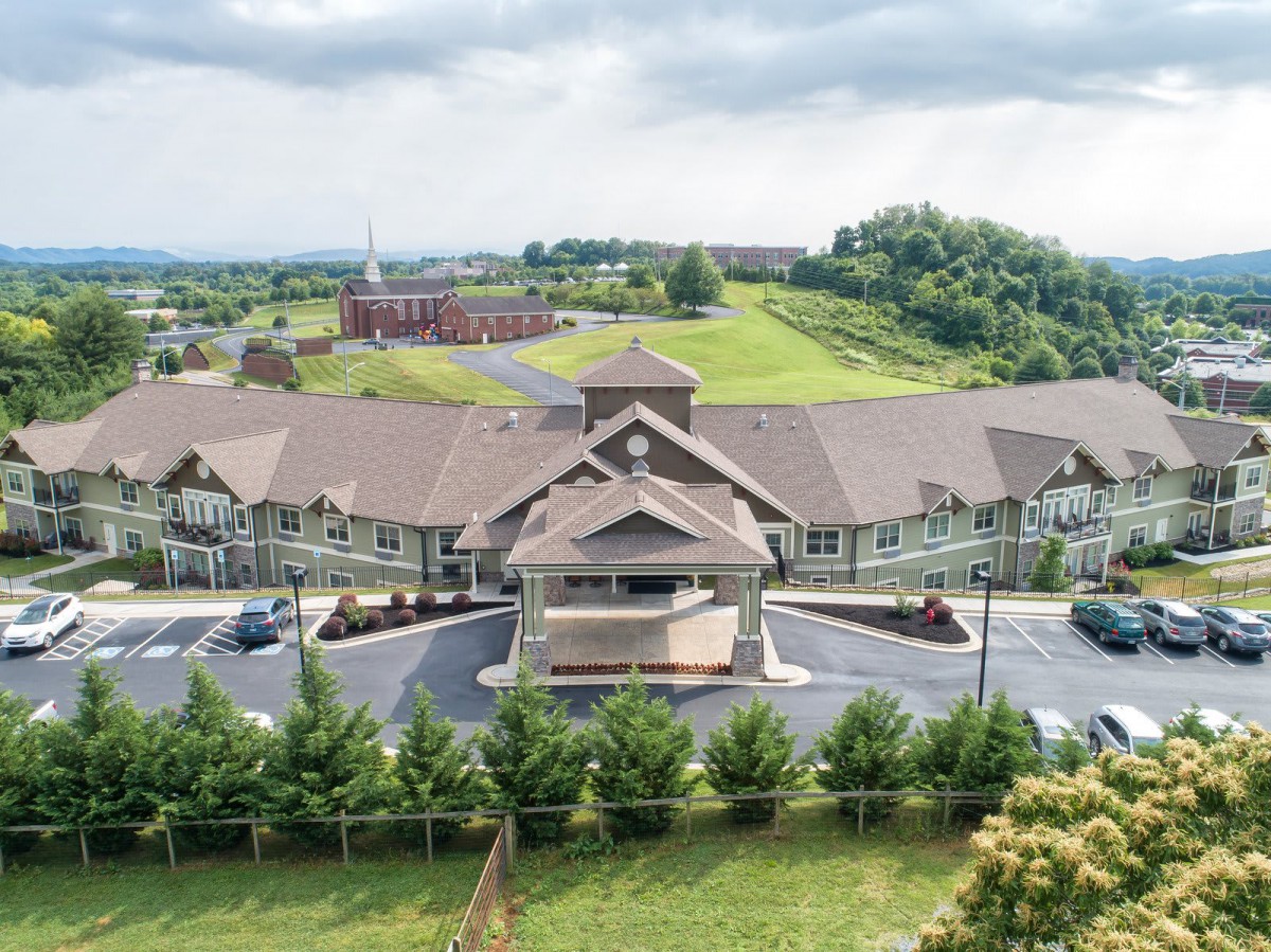 Aerial view of a large building surrounded by greenery and parking areas under a cloudy sky.