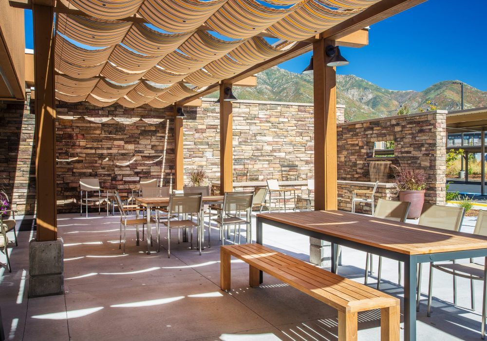A spacious outdoor patio with wooden furniture and a pergola for shade, surrounded by stone walls on a sunny day.