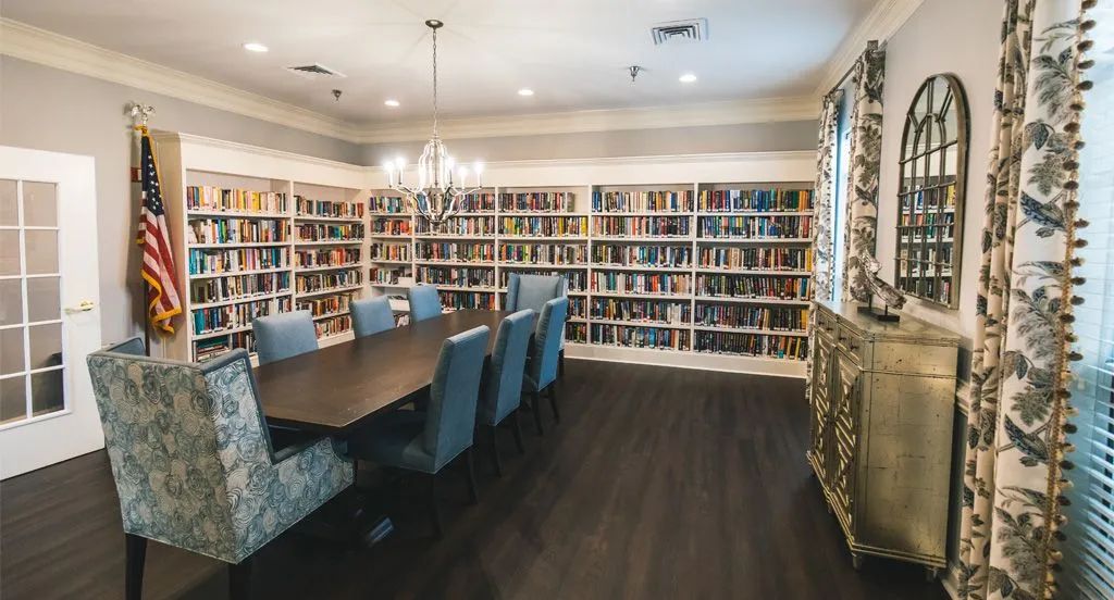 A cozy library room with a wooden table surrounded by chairs and shelves filled with books.