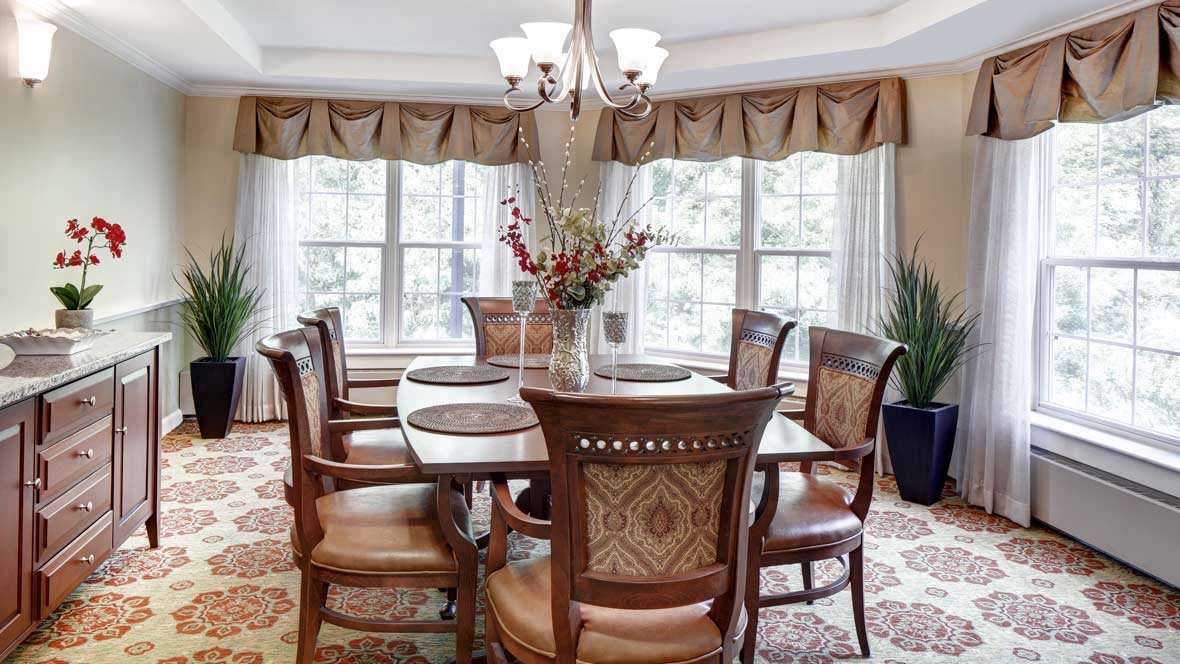 A well-appointed dining room with a large table, elegant chairs, and a chandelier, surrounded by windows with curtains.