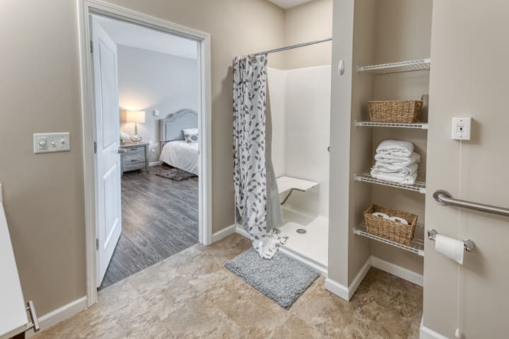 A modern bathroom with a shower and neatly organized shelves, leading into a cozy bedroom.