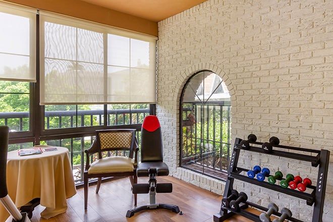A cozy home gym with exercise equipment, including a weight bench and dumbbells, is brightened by natural light filtering through the blinds.