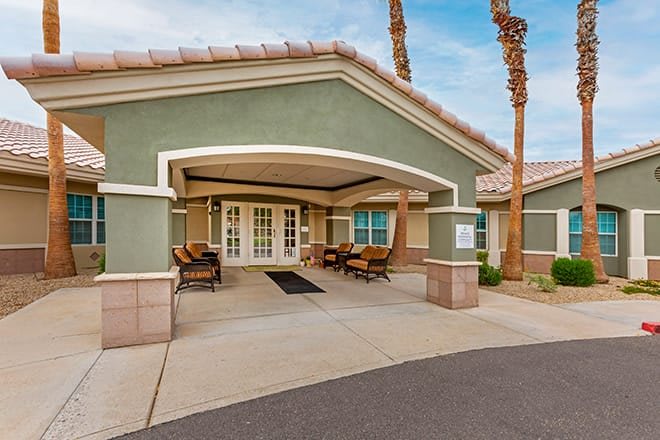 A welcoming building entrance featuring a covered patio with seating and palm trees in the background.