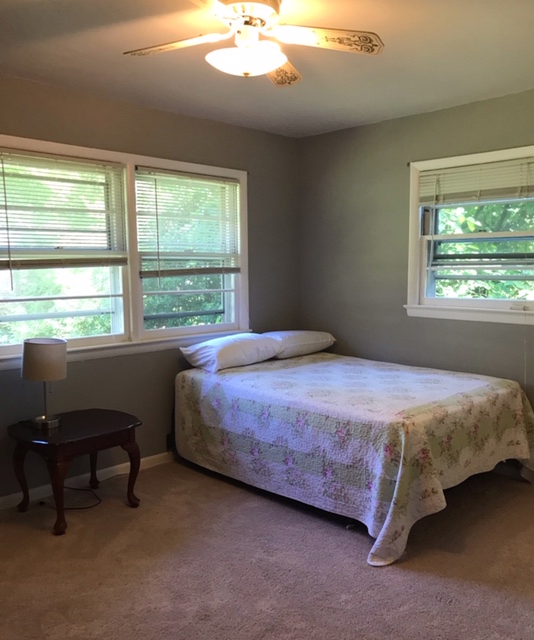A minimalist bedroom with a bed, chair, and ample natural light from two windows.