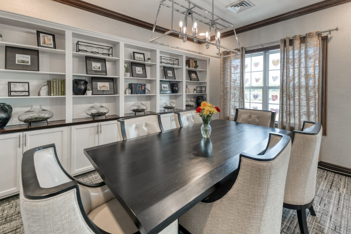 A modern dining room features a large dark table surrounded by upholstered chairs, with bookshelves in the background and a stylish chandelier overhead.