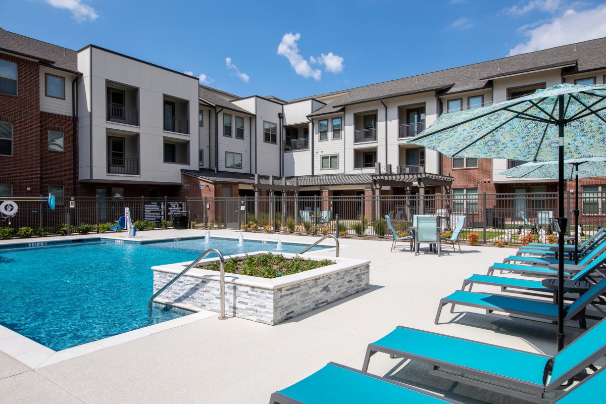 A modern apartment complex featuring an inviting outdoor swimming pool surrounded by lounge chairs on a sunny day.
